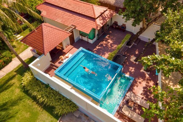 Over udsigt over en resorts swimmingpool med klart blåt vand, omgivet af liggestole og palmer.