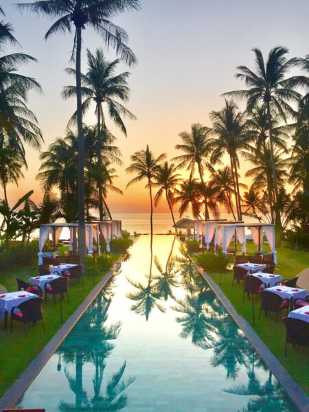Pool med borde og stole ved Kupu Kupu Phangan Beach Villas ved solnedgang omgivet af palmetræer