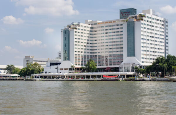 Stor hvid bygning ved vandkant ligner Wyndham Bangkok Menam Riverside eller Ramada Plaza