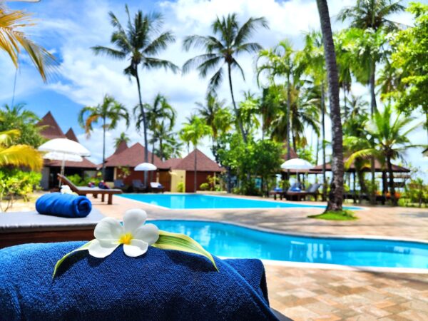 Swimmingpool ved Sivalai Beach Resort omgivet af palmer og loungestole, blå himmel og hvid blomst på håndklæde i forgrunden.