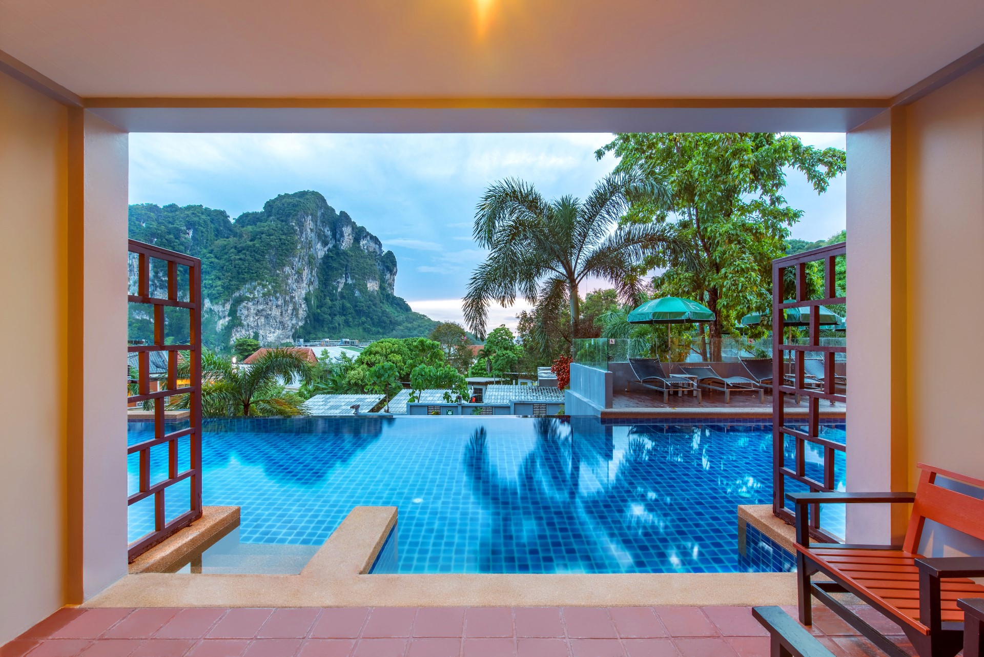 Udsigt fra overdækket terrasse på Cha Da Resort til infinity pool, palmer og bjerglandskab i solskin i Krabi.