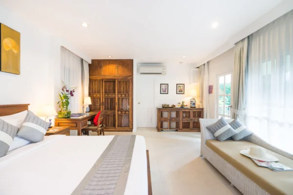 Lyst soveværelse på Laksasubha Hua Hin Hotel med stor seng, træmøbler og sollys gennem lette gardiner.