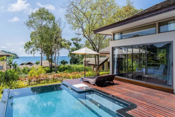 Moderne villa med glaspartier, træterrasse og infinity pool med udsigt over frodig natur og have på eksotisk stranddestination.
