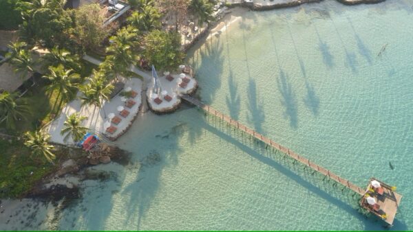 Træmole strækker sig over klart turkisblåt hav ved sandstrand med palmer og solsenge på Beach Natural Resort i Thailand.